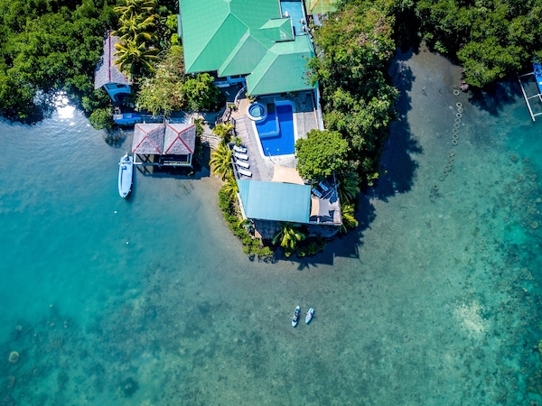 Luftbild einer tropischen Inselanlage mit Pool, Bootsanleger und türkisfarbenem Wasser.