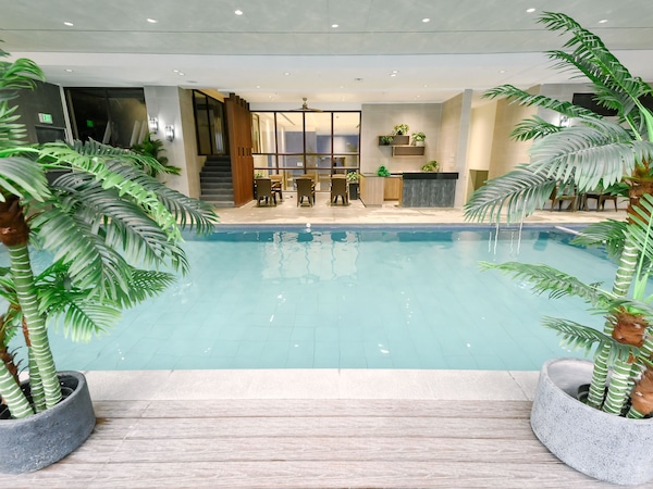 Hallenbad mit Holzdeck, Palmen in Kübeln und einem Sitzbereich mit Tisch und Stühlen.